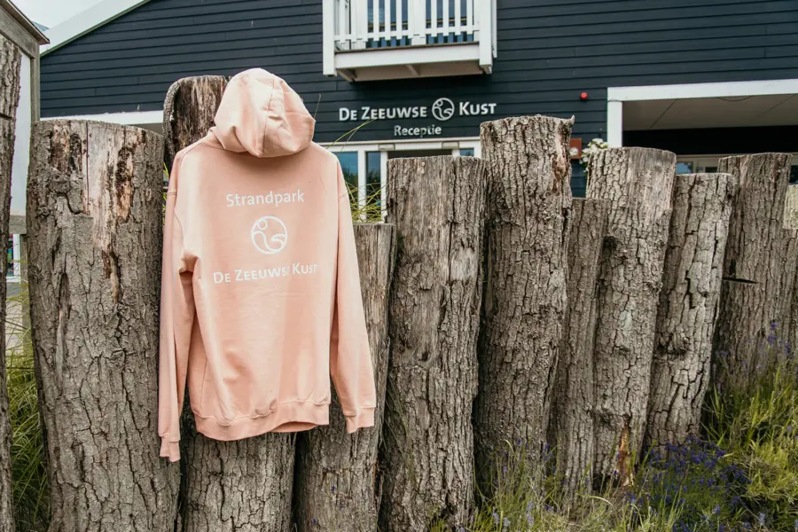 Zachte roze hoodie met logo van Strandpark De Zeeuwse Kust hangt aan een houten schutting bij de receptie