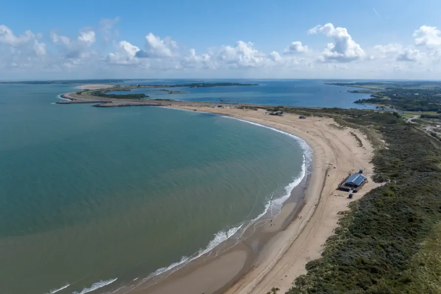 Luchtfoto van het uitgestrekte zandstrand bij Strandpark De Zeeuwse Kust met helderblauw water duinen en een strandpaviljoen aan de kust